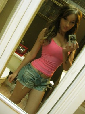 amateur photo This brunette girl, taking a selfie in the bathroom, is hot and has nice cleavage. She has blue nails, a tattoo, and is wearing a pink shirt and jean 